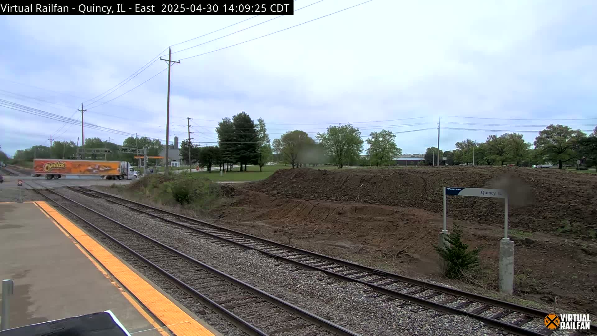 A train platform with an orange tactile strip next to train tracks, a large pile of dirt nearby, and a Cheetos-branded semi-truck crossing a road in the background under a mostly cloudy sky.