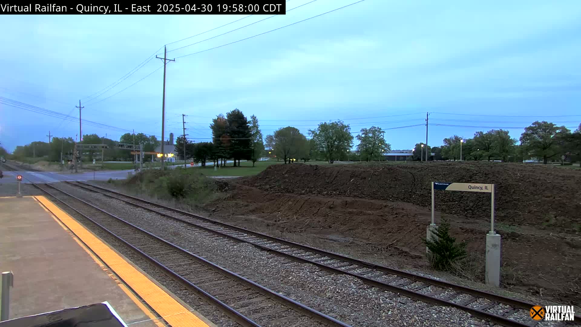 A train platform with two sets of tracks extends into the dusk, alongside a freshly excavated earthen berm and a few trees under a twilight sky.