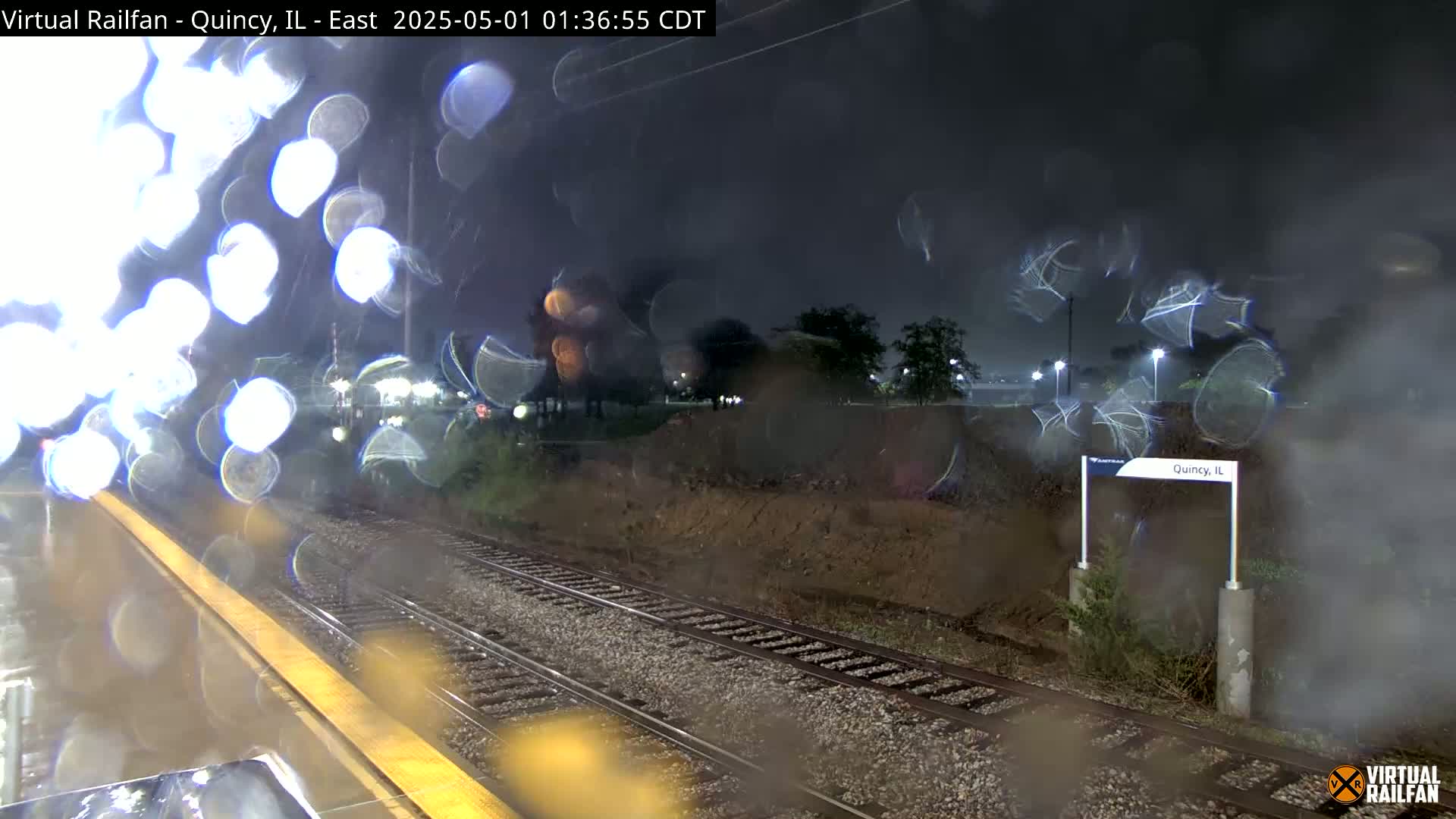 A nighttime view through rain-streaked glass shows train tracks and a station platform, with blurry lights and vegetation visible in the background.