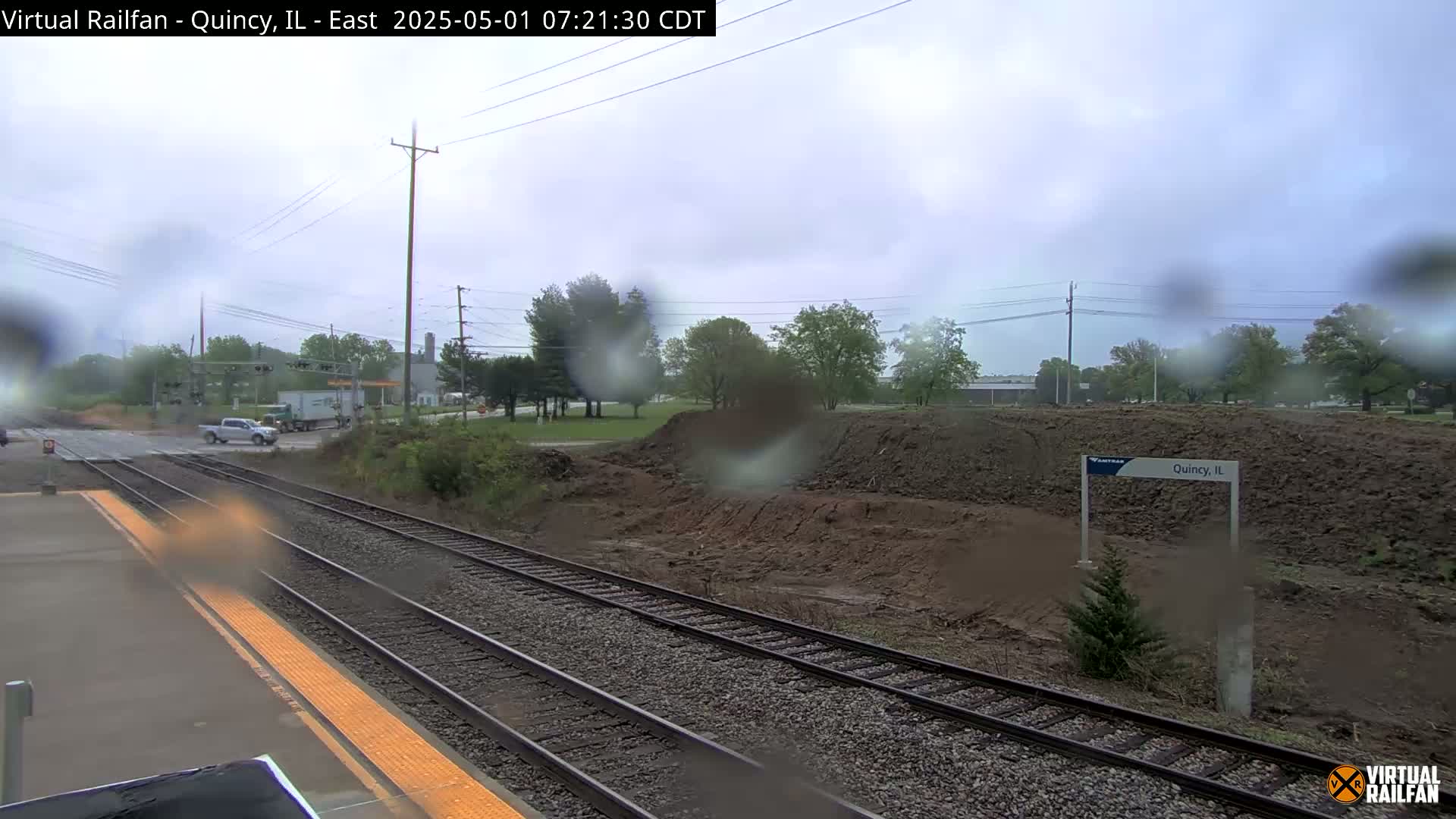 A rain-streaked view of a train platform shows railroad tracks, a road crossing, and an area of excavated earth, with a few trees and vehicles visible in the background.