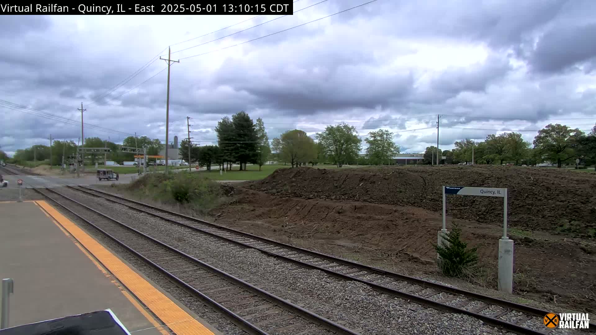 A train station platform with a view of train tracks, a large pile of dirt, and a partly cloudy sky.