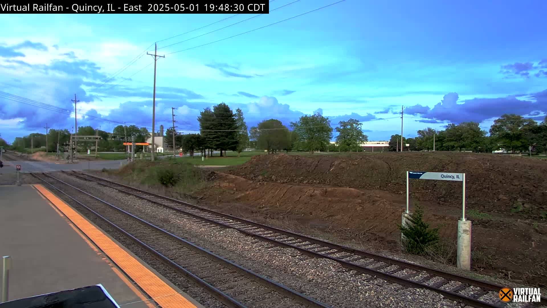 A train platform with multiple tracks under a partly cloudy evening sky overlooks a large pile of earth.