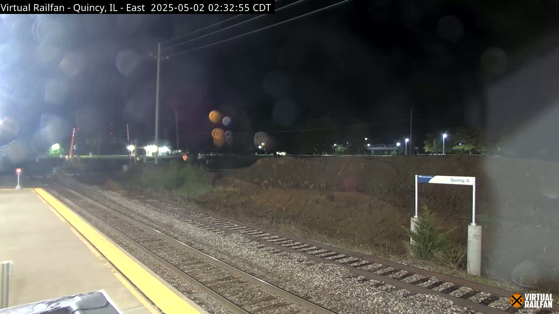 A nighttime view of train tracks at a station, with a dirt embankment beyond the platform and some light rain or mist in the air.