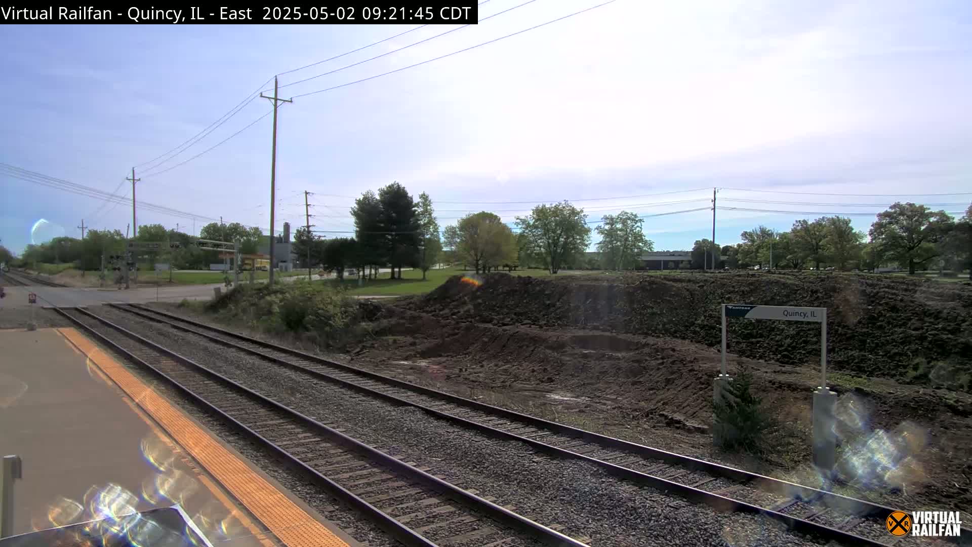 A train platform with multiple tracks runs alongside a pile of dirt, under a partly cloudy sky.