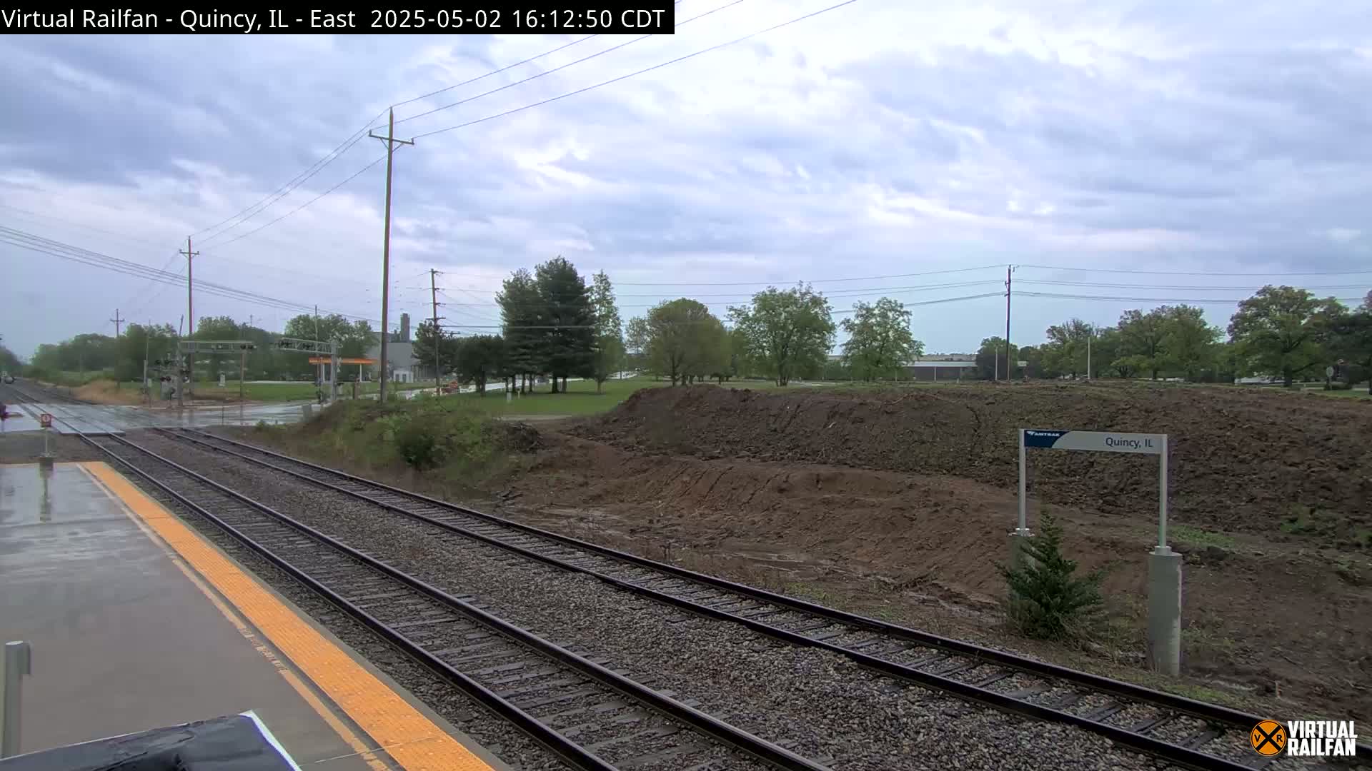 A wet train platform overlooks a section of railway tracks and a large pile of earth under an overcast sky.