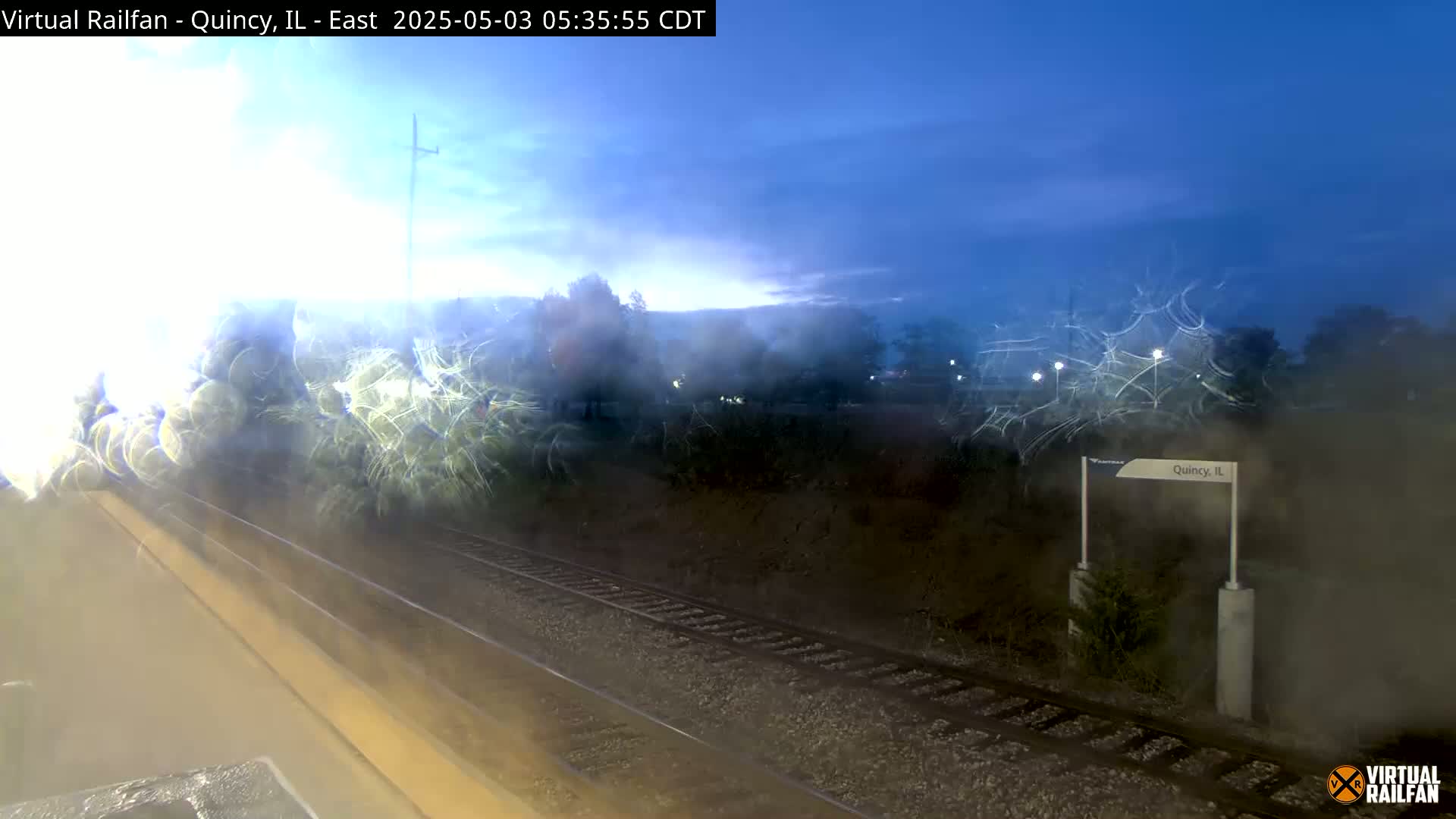 A view from a train window shows a train station at dawn, with a sign indicating Quincy, IL, and train tracks stretching into the distance.