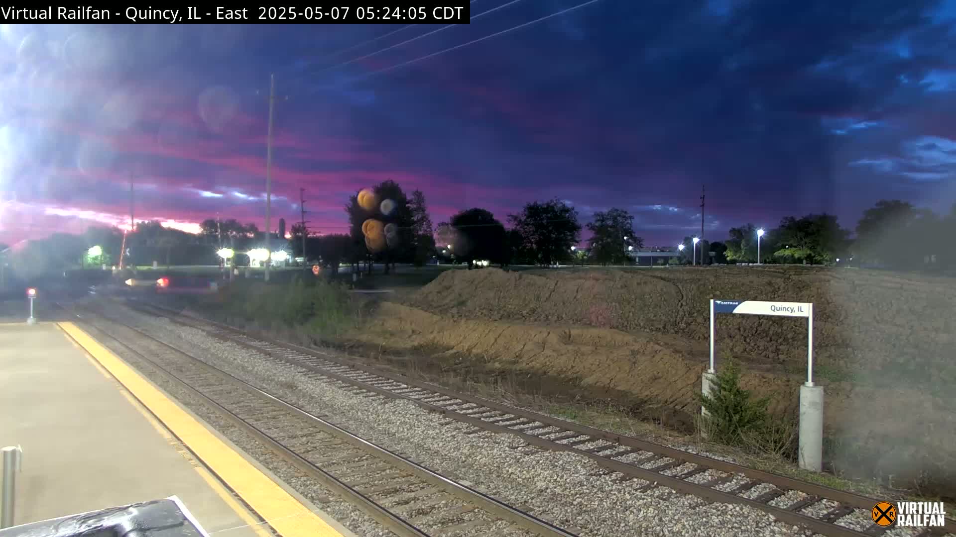 A train station platform with railroad tracks under a twilight sky displaying vibrant purple and pink hues.