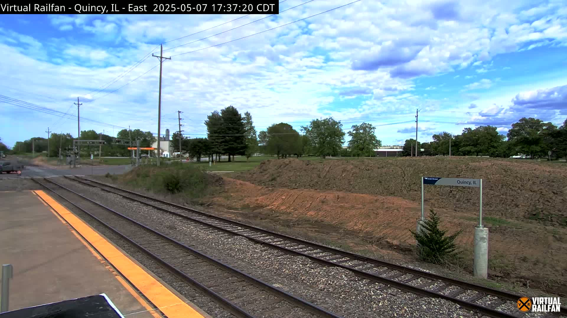 A mostly sunny day with partly cloudy skies shows a train station platform next to a railway with a dirt embankment and trees in the background.