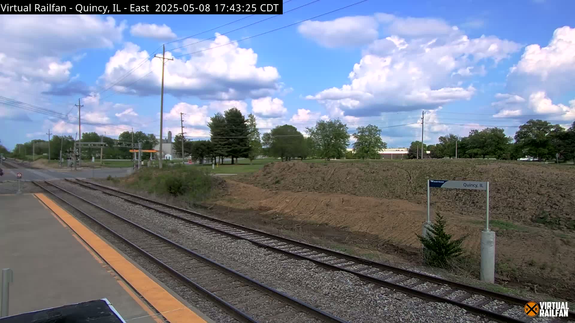 A view of train tracks under a partly cloudy sky, with a grassy area and trees in the background.