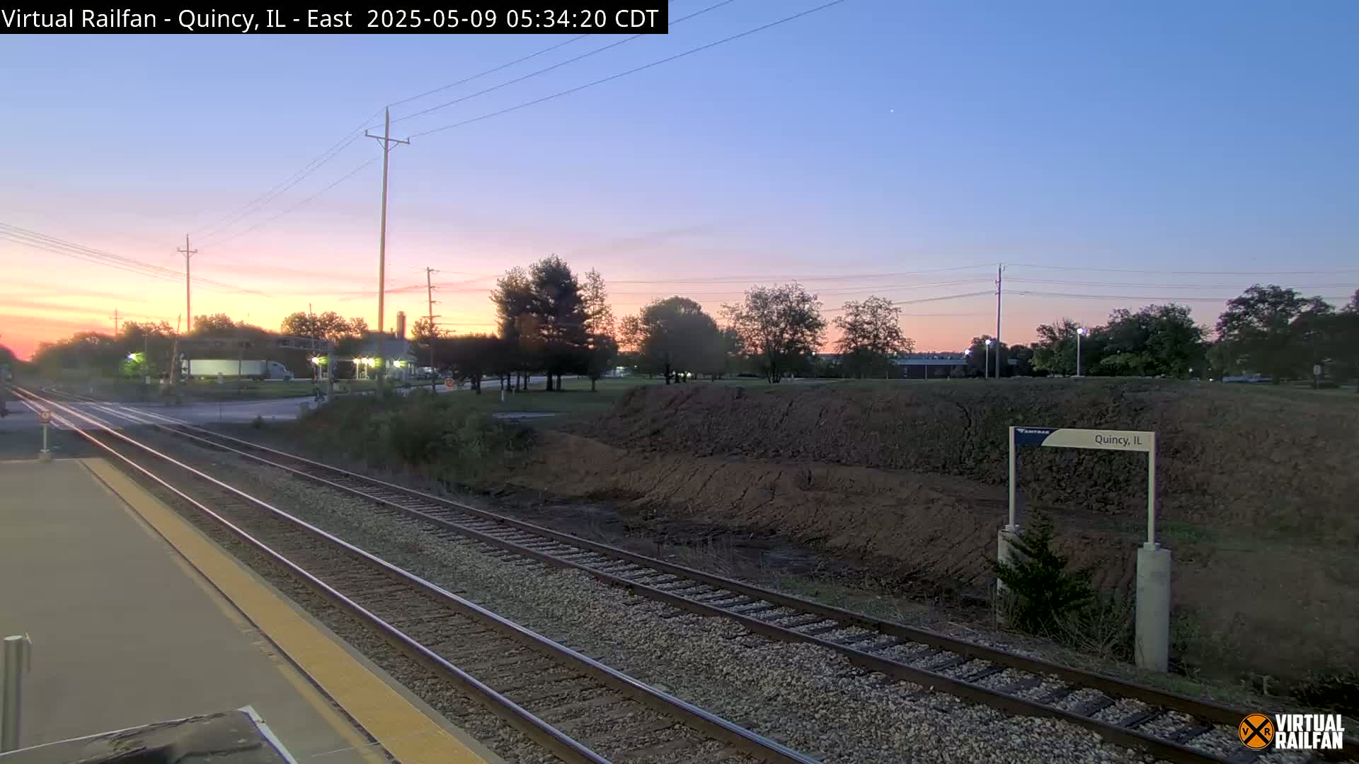A train station platform at sunrise, with two sets of train tracks extending into the distance past a low dirt embankment.