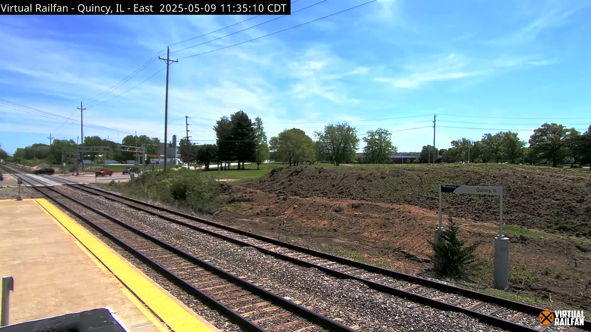 A train station platform with two train tracks stretches into the distance under a mostly sunny sky, past a patch of exposed earth and some trees.