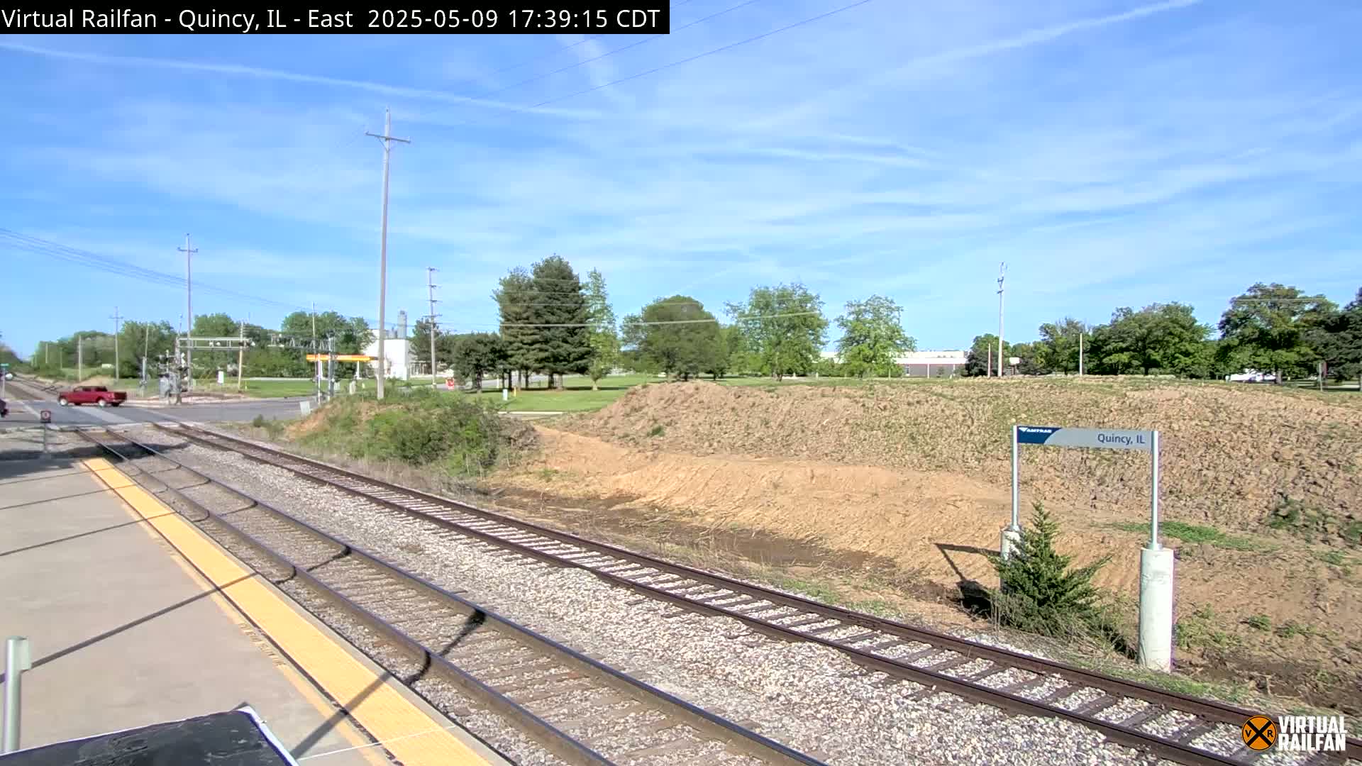 A train track runs through a mostly sunny landscape with a red pickup truck visible in the distance, and a small patch of bare earth next to it.