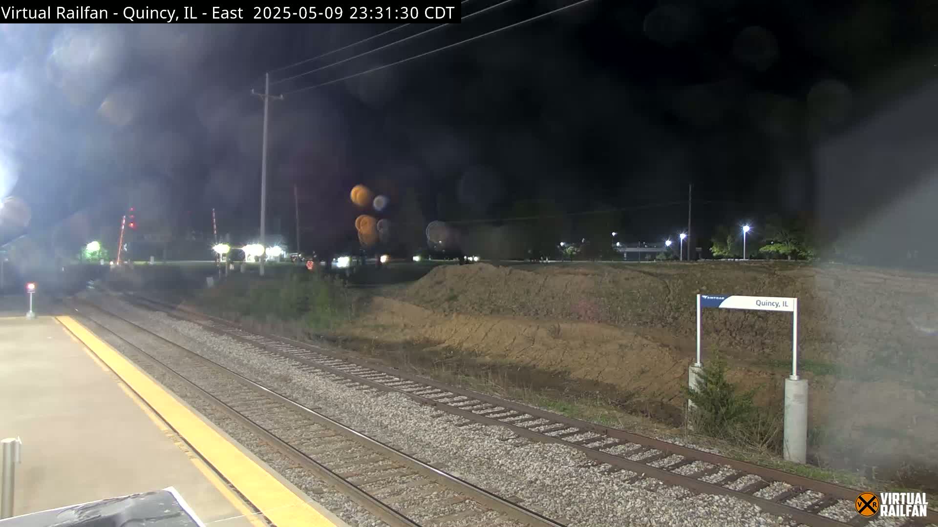 A nighttime view of train tracks at a station, with a dirt embankment visible beyond the tracks, under hazy conditions.