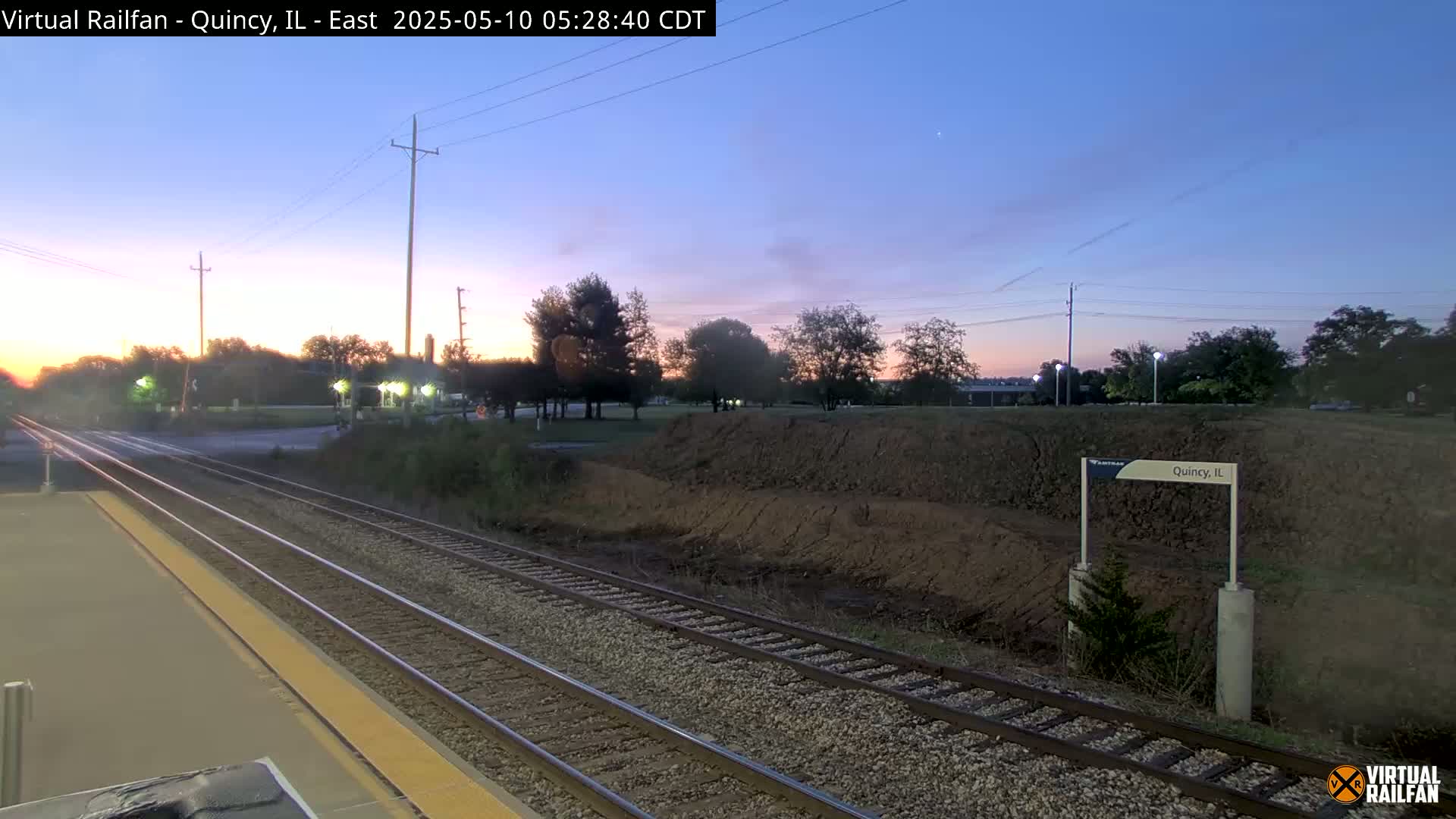 A sunrise view of train tracks at a station, with a low earthen embankment beyond.