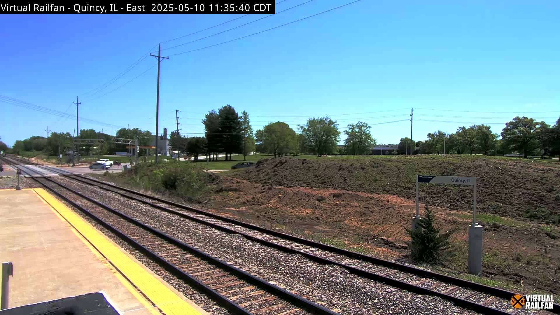 A sunny day at a train station, with railroad tracks extending into the distance, past a patch of disturbed earth and some trees, and a road crossing in the background.