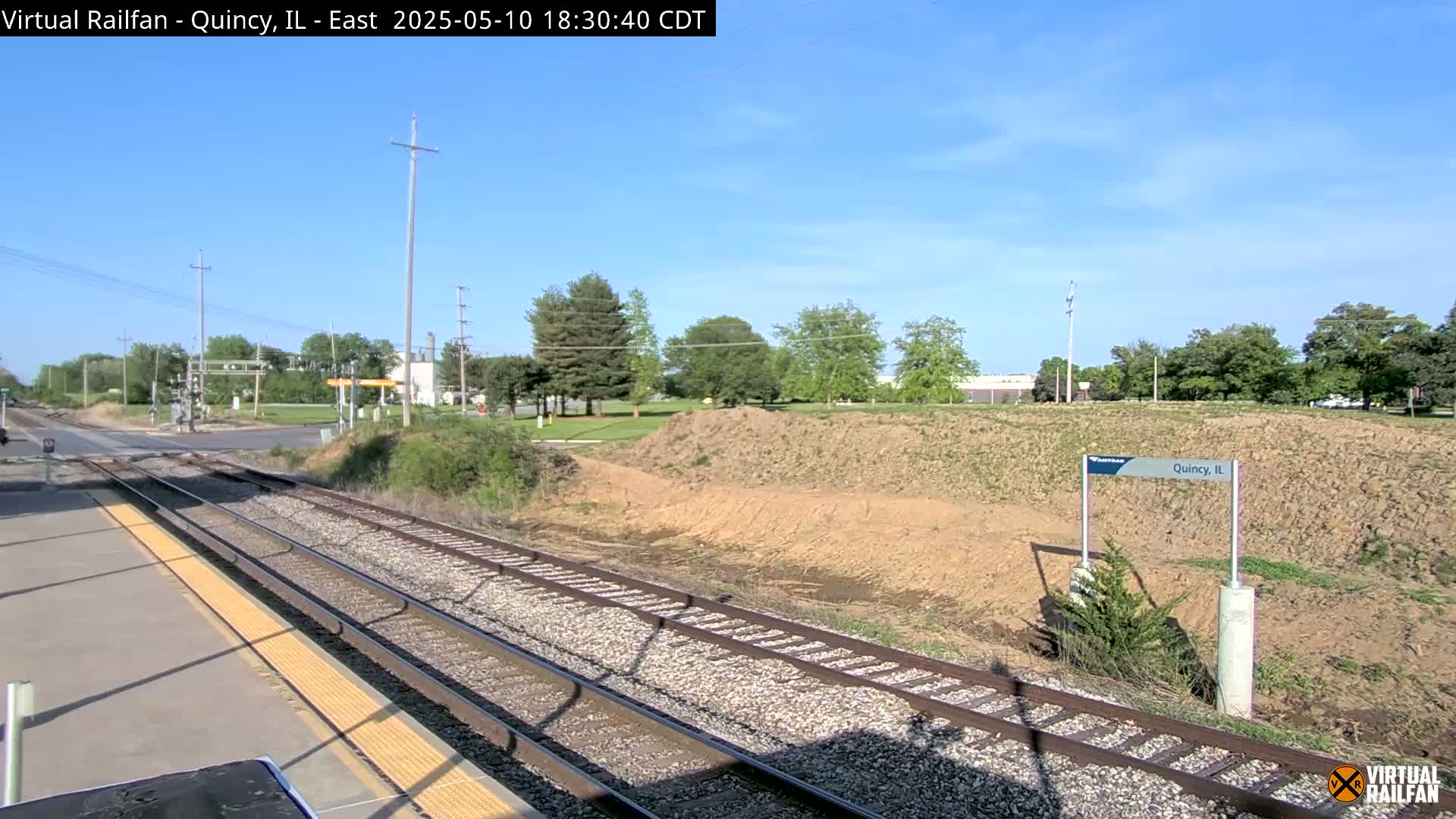 A train platform overlooks a railway track that runs alongside a dirt embankment, under a mostly clear sky.