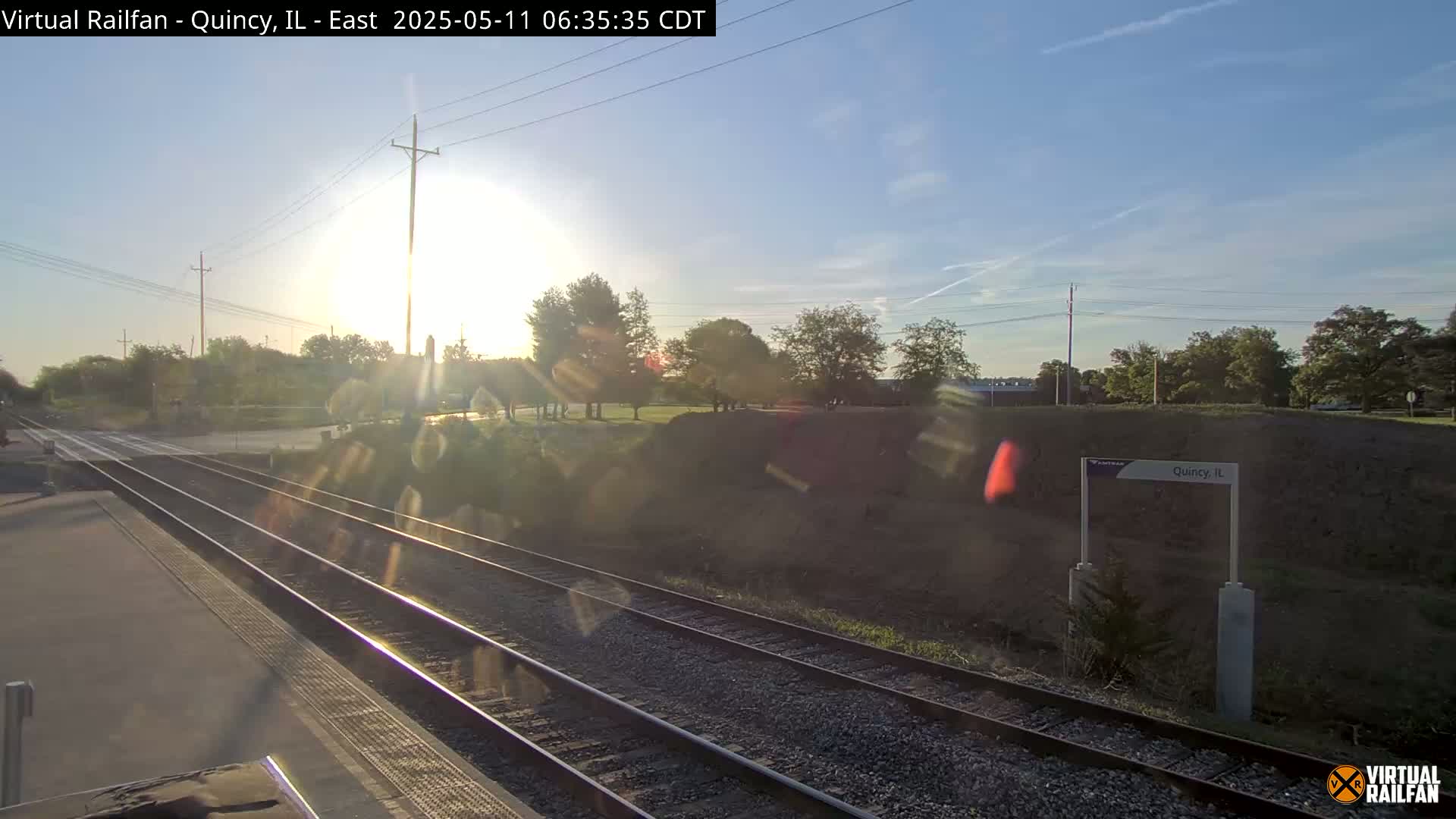 A sunny morning view of train tracks at a station, with trees and a grassy embankment in the background.