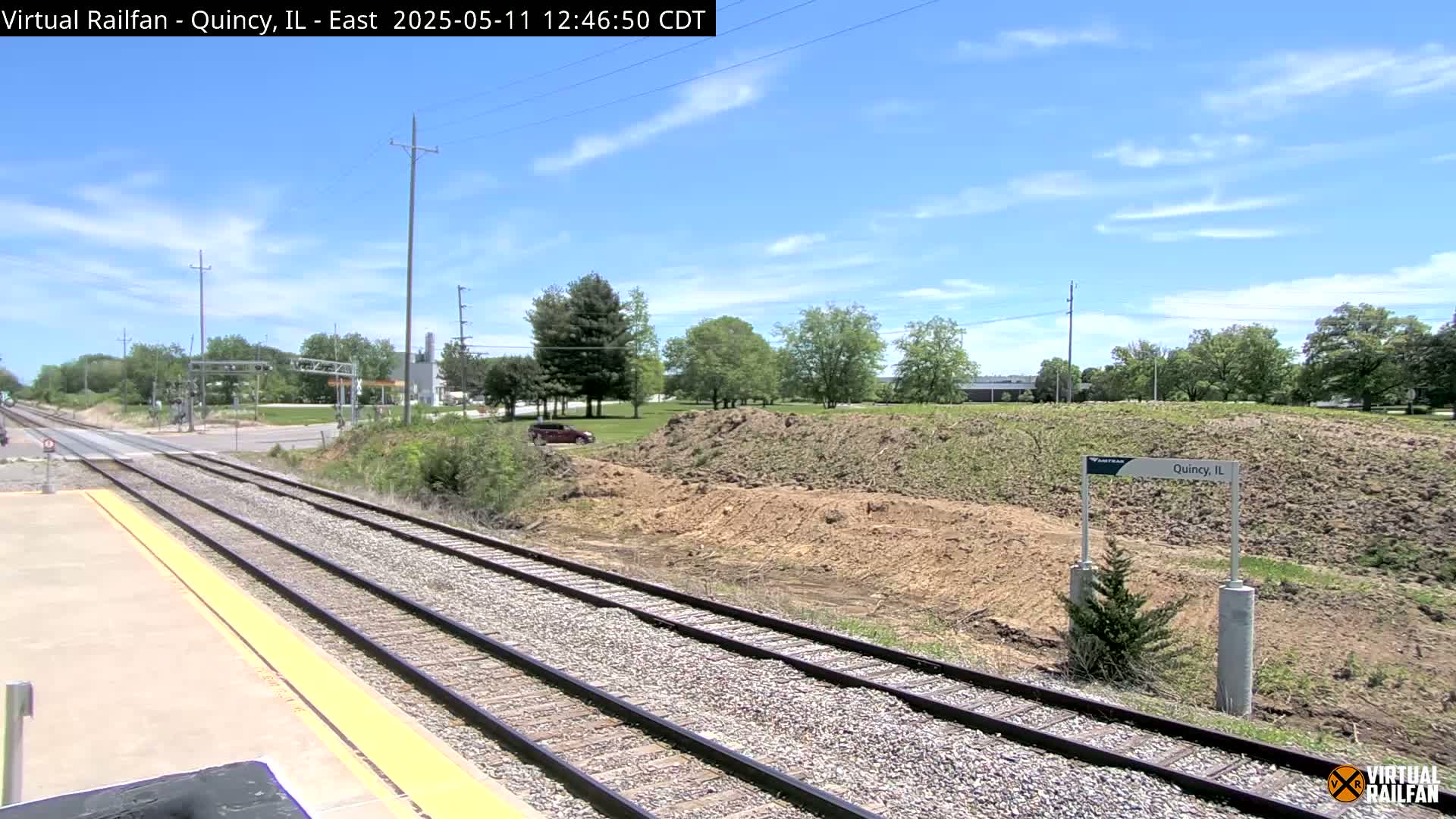 A sunny day reveals a train track splitting into two lines, passing beside a cleared, earthy mound with sparse vegetation, and a roadway visible in the distance.