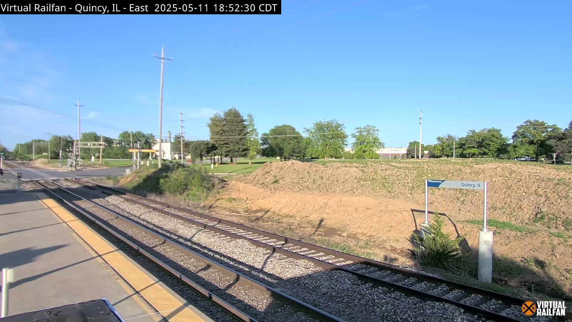 A train platform overlooks a train track leading to a road crossing under a clear blue sky, with green trees and a bare, tan hill in the background.
