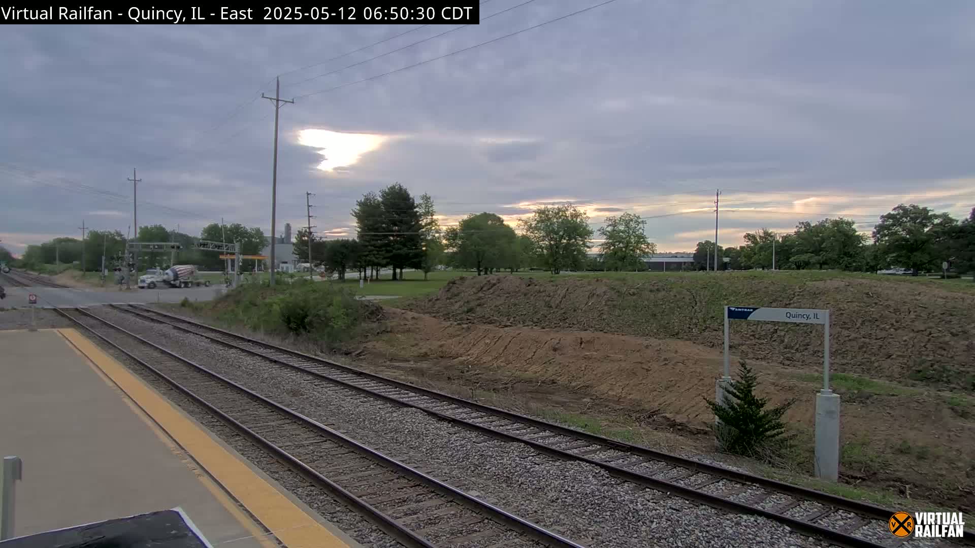 A mostly cloudy sunrise illuminates a train station platform with several train tracks, a road crossing, and a cement mixer in the distance.