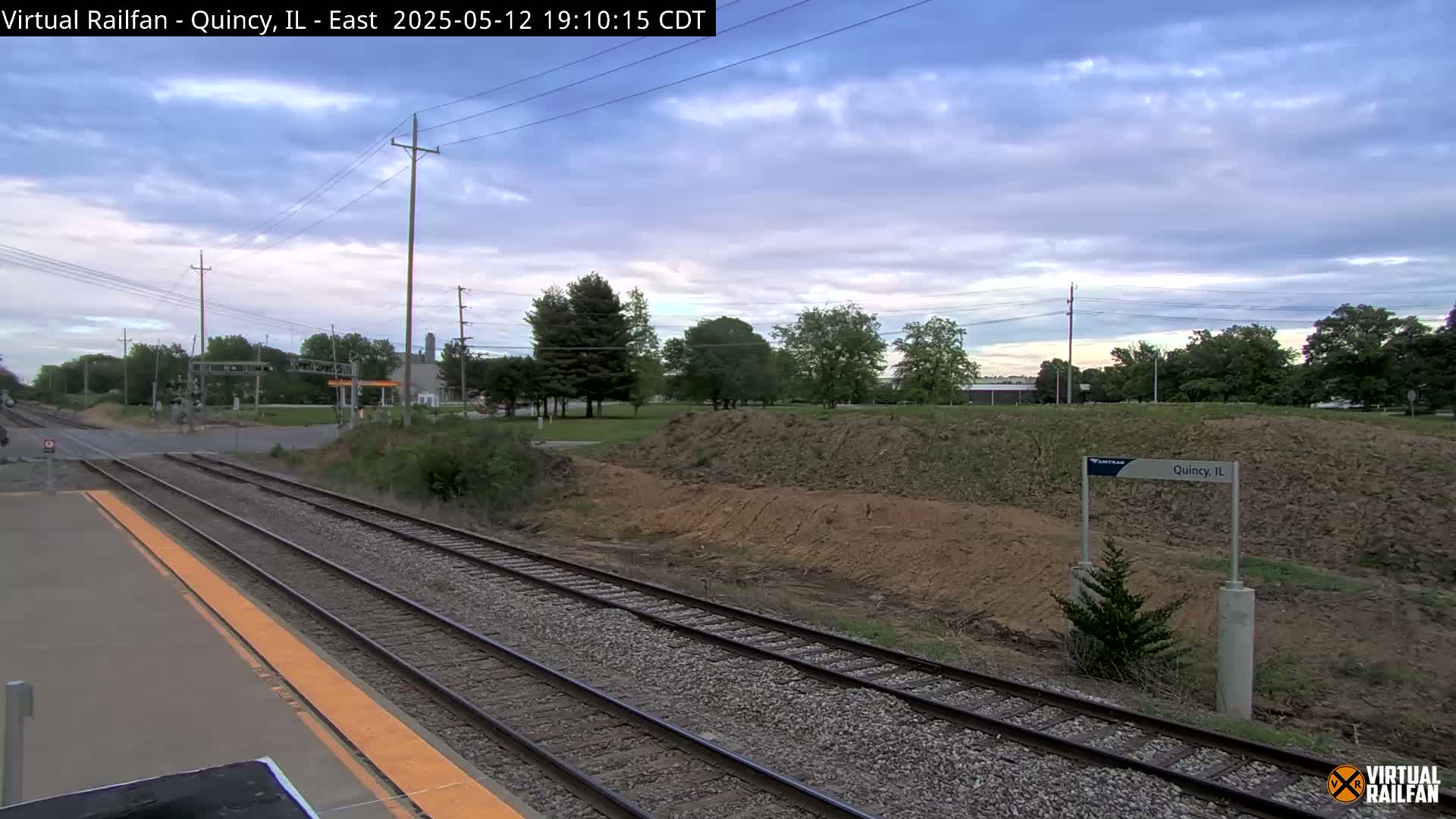 A train platform with multiple train tracks stretches into the distance under a partly cloudy sky.