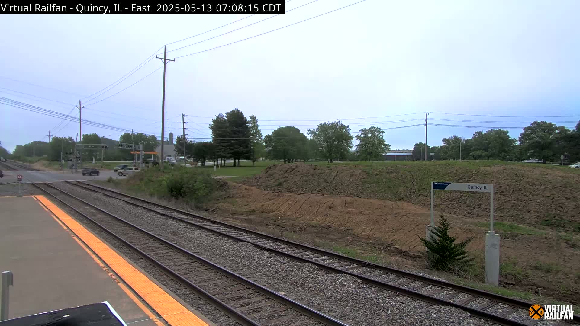 A train platform with two sets of tracks is shown on an overcast day, with a pile of dirt and trees visible beyond the tracks.