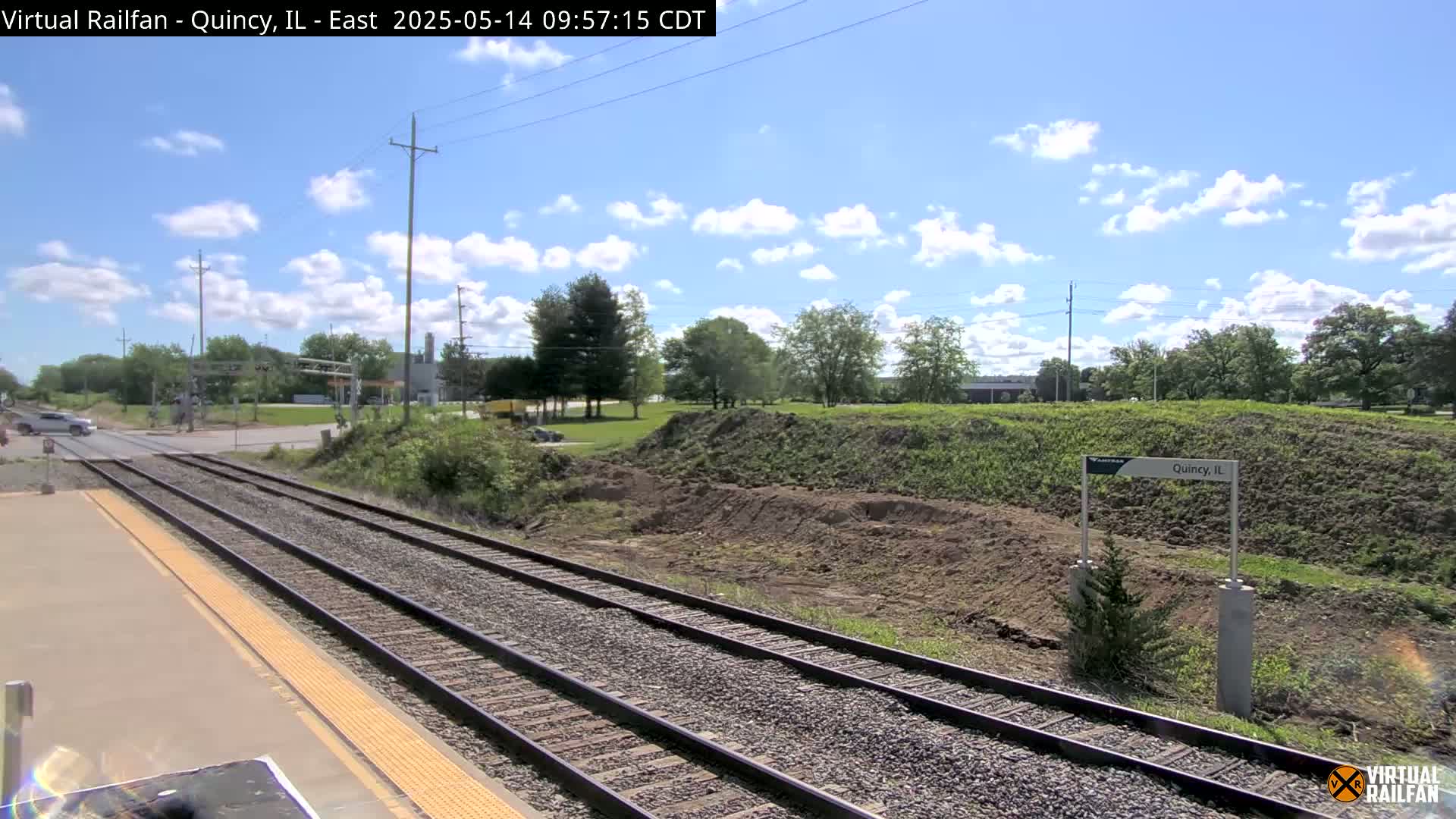 A train platform with two sets of tracks stretches into a sunny landscape under a partly cloudy sky,  beyond which is a mound of freshly turned earth and some greenery.