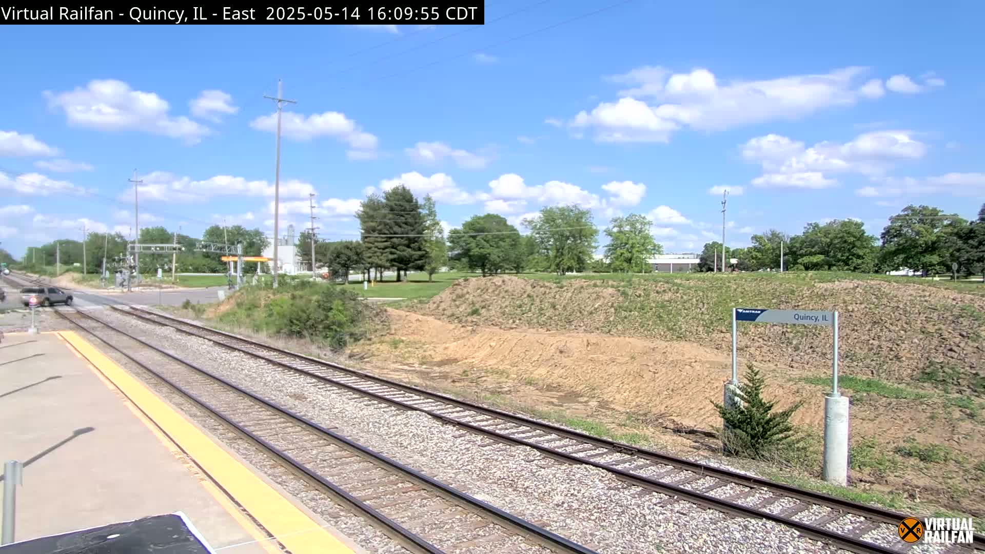 A partly sunny day reveals train tracks leading to a station, with a grassy hill and trees in the background.