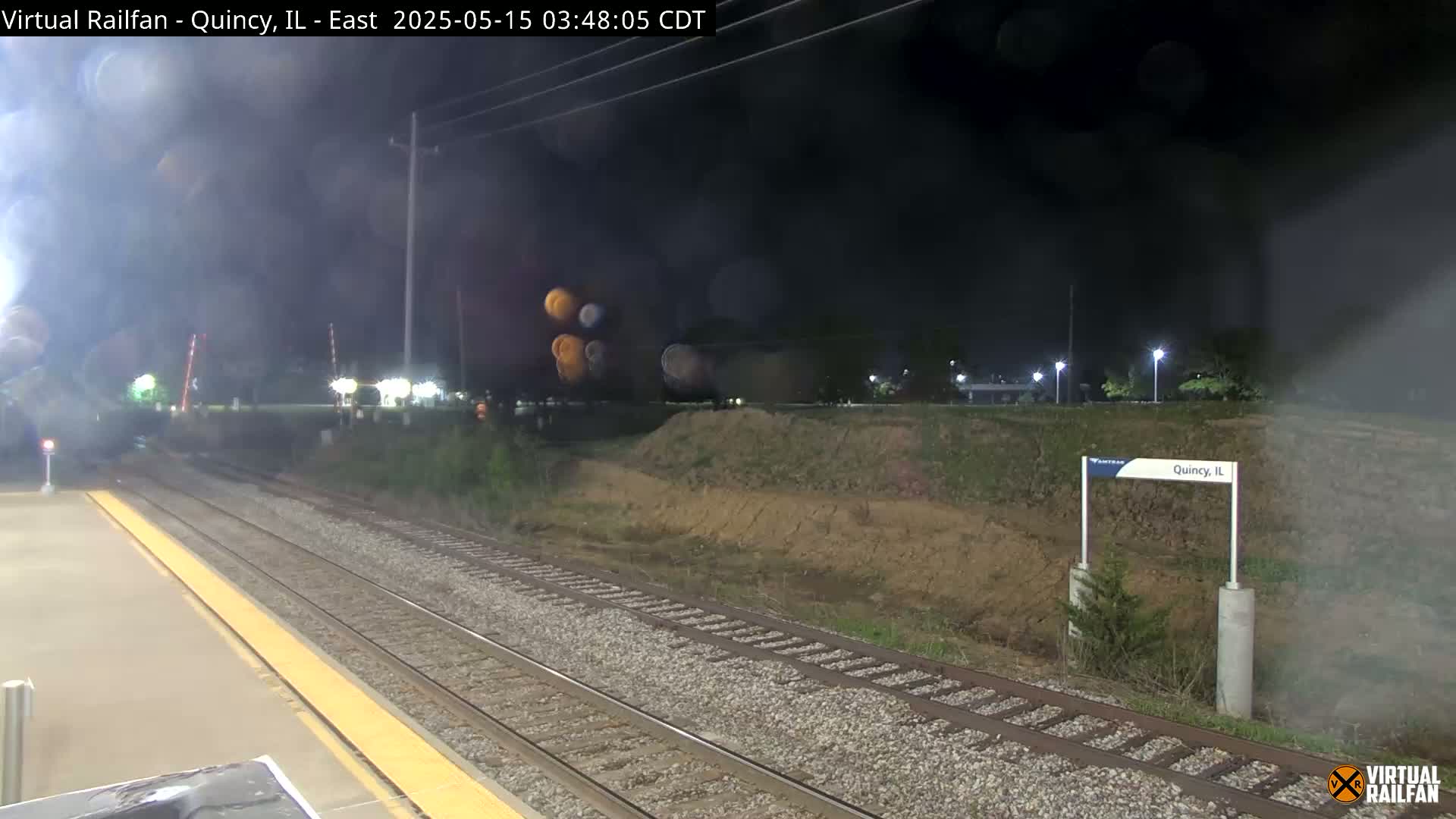 At night, a train station platform is seen with train tracks extending into the distance, under hazy conditions.