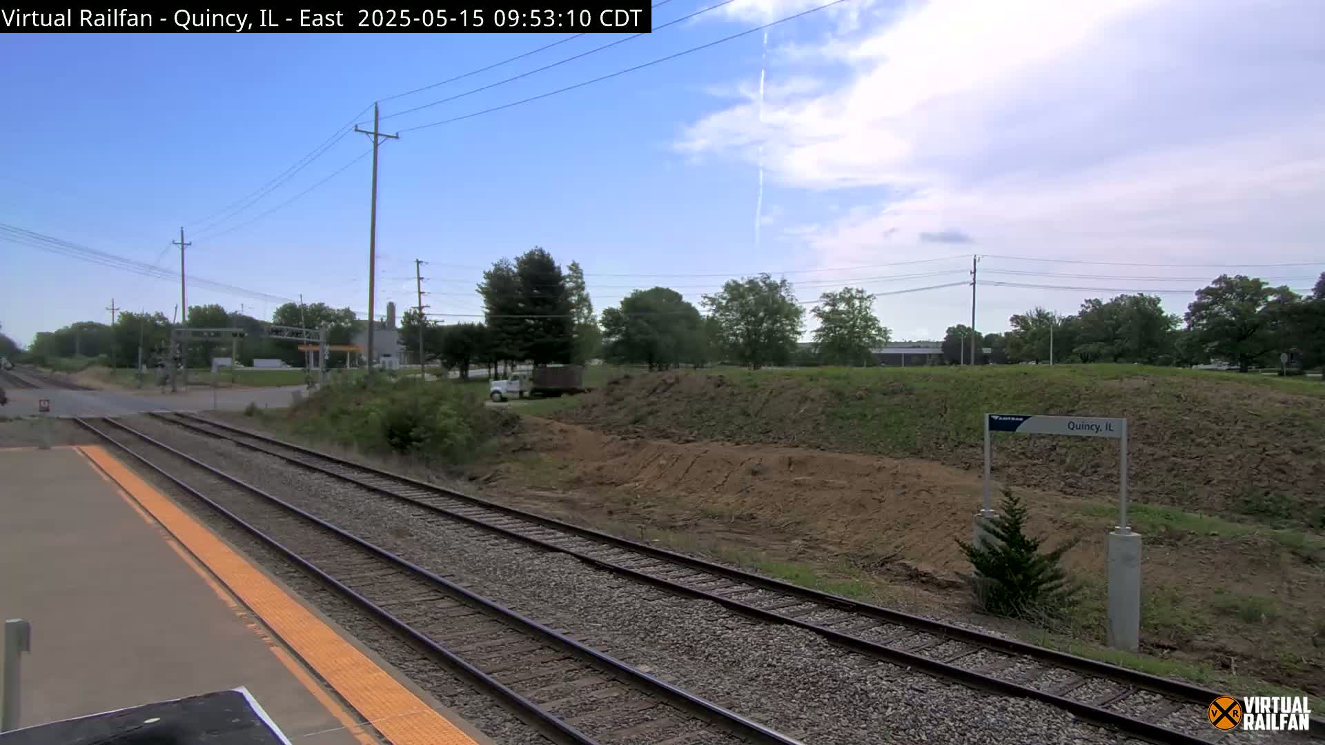 A train platform with two sets of tracks runs alongside a dirt embankment under a partly cloudy sky.