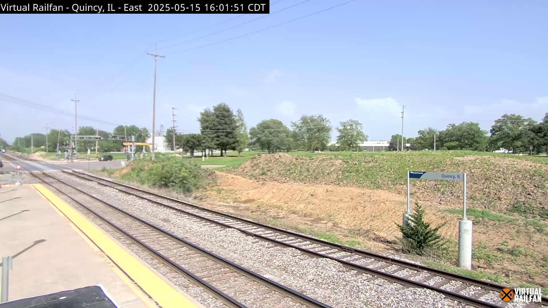 A train station platform overlooks a section of train tracks that lead toward a road crossing, under a mostly clear sky.