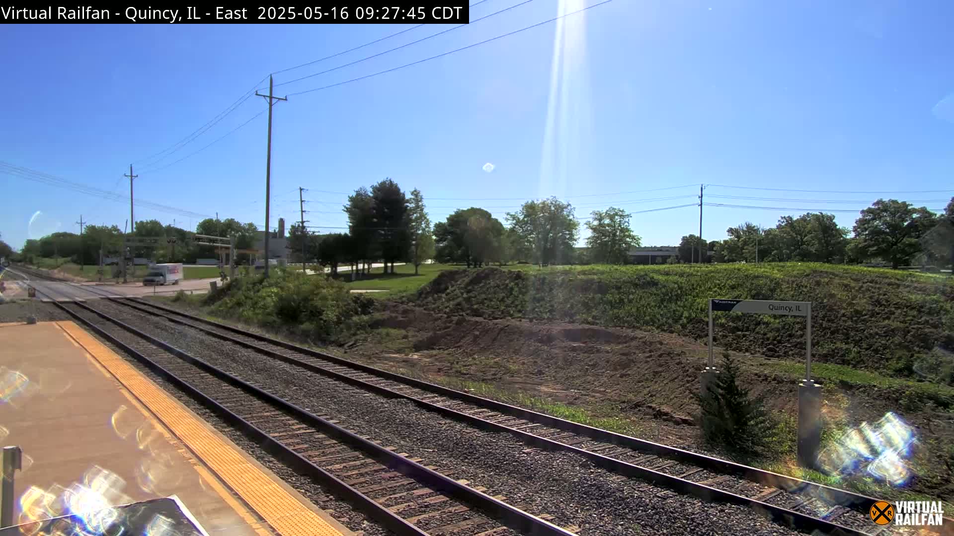 A sunny day at a train station with two sets of train tracks, a road crossing in the distance, and a grassy embankment.