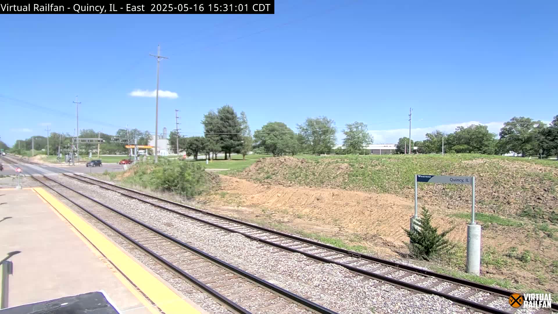 A sunny day at a train station, showing train tracks extending into the distance past a small hill and some sparse vegetation.