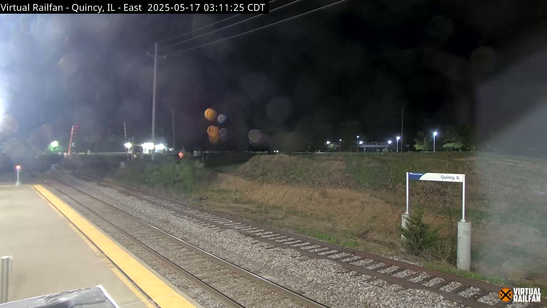 A nighttime view of train tracks at a station, possibly during light rain or mist.