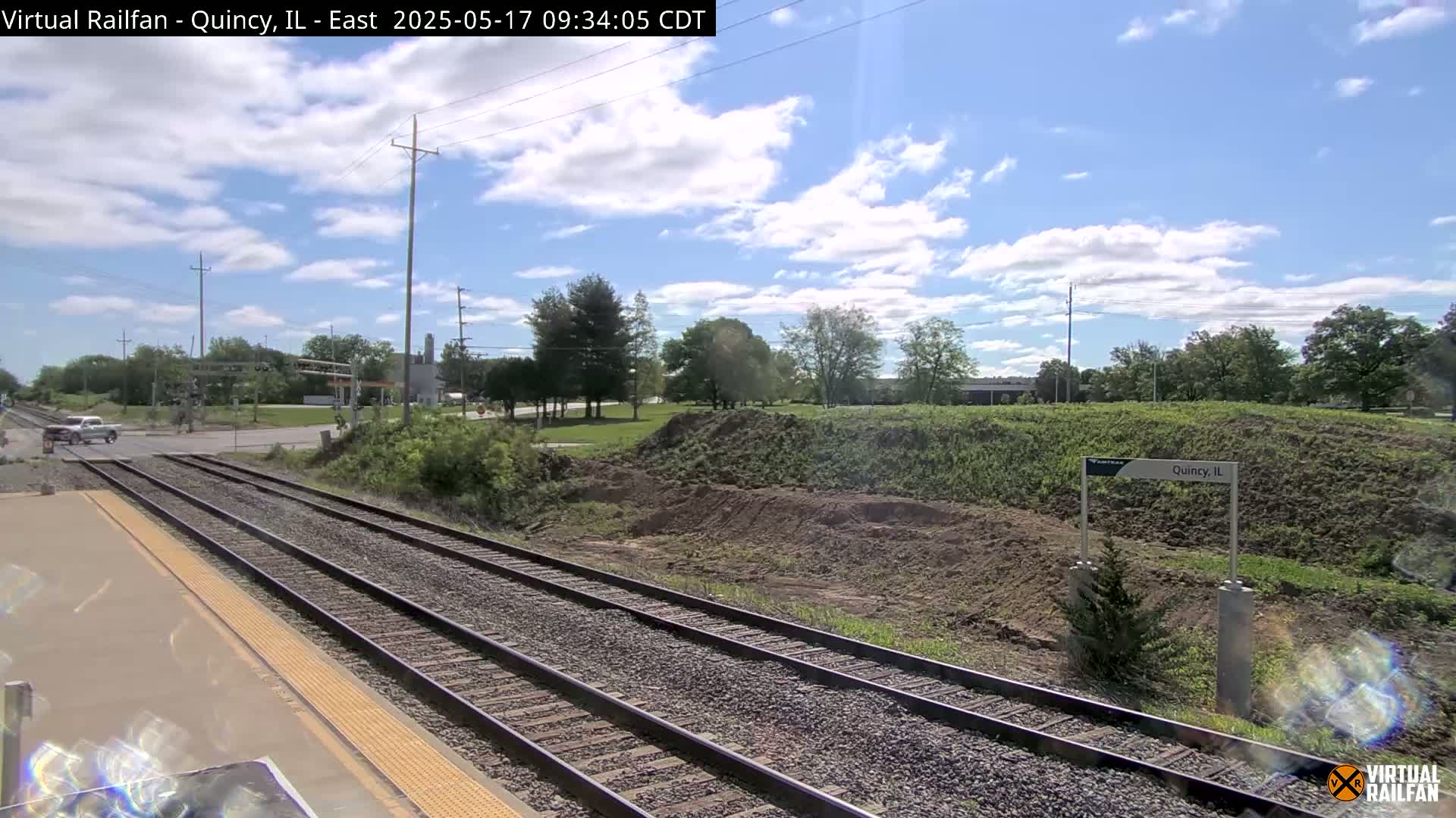 A view from a train platform shows several train tracks leading toward a road intersection under a partly cloudy sky.