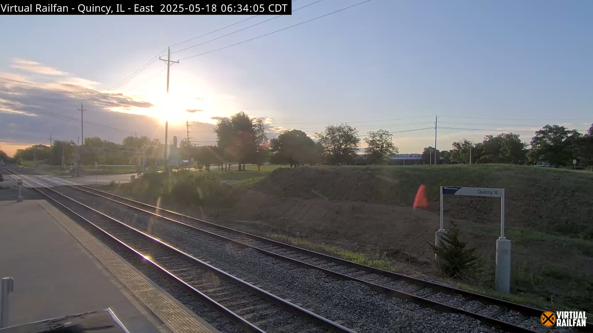 A sunrise illuminates a train platform with two sets of tracks and a grassy area beyond, under a mostly clear sky.