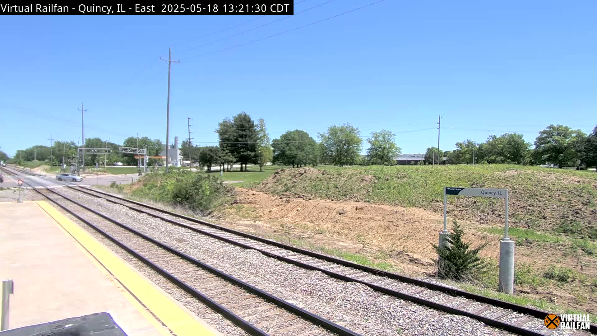A train track stretches into the distance on a sunny day, past a grassy area with some bare earth and a few trees.