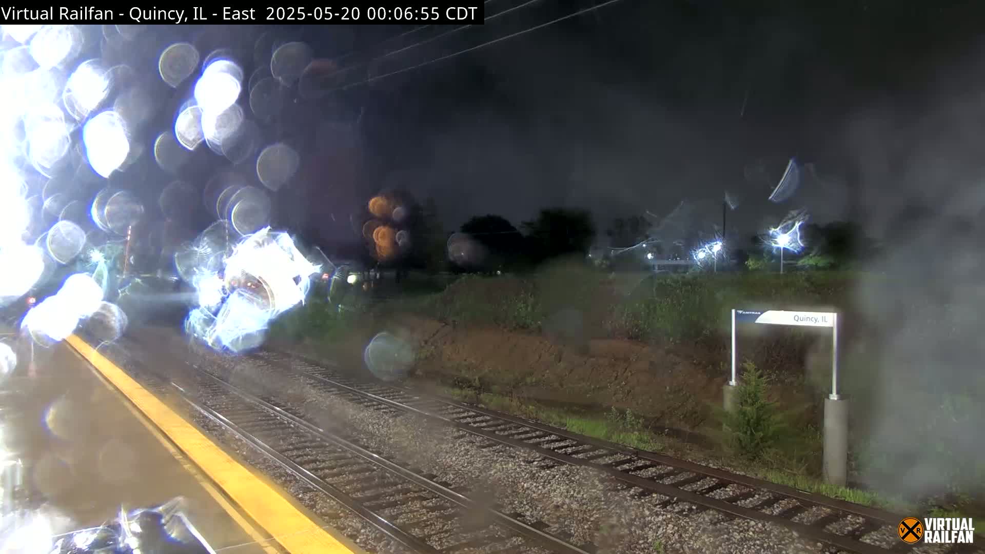 A rain-streaked nighttime view from a train shows a railroad platform and tracks leading to a station sign in Quincy, IL.