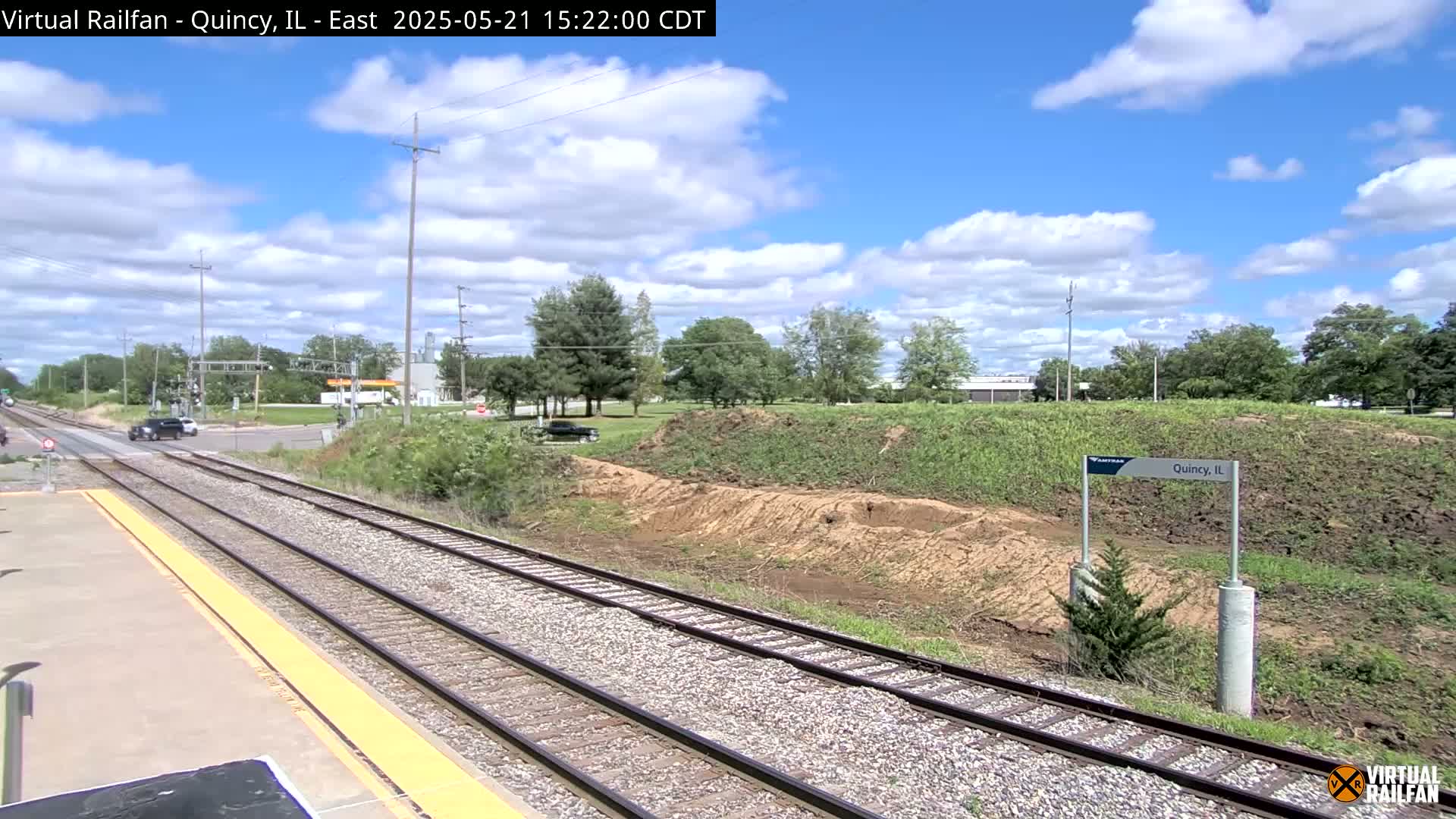 A train track at a station with a partly cloudy sky, some cars on a road crossing the tracks, and some earthworks alongside the tracks.