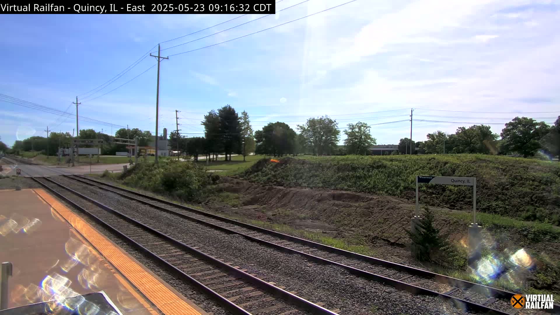 A train station platform with two sets of train tracks, under a partly cloudy sky, and surrounded by greenery and a dirt embankment.