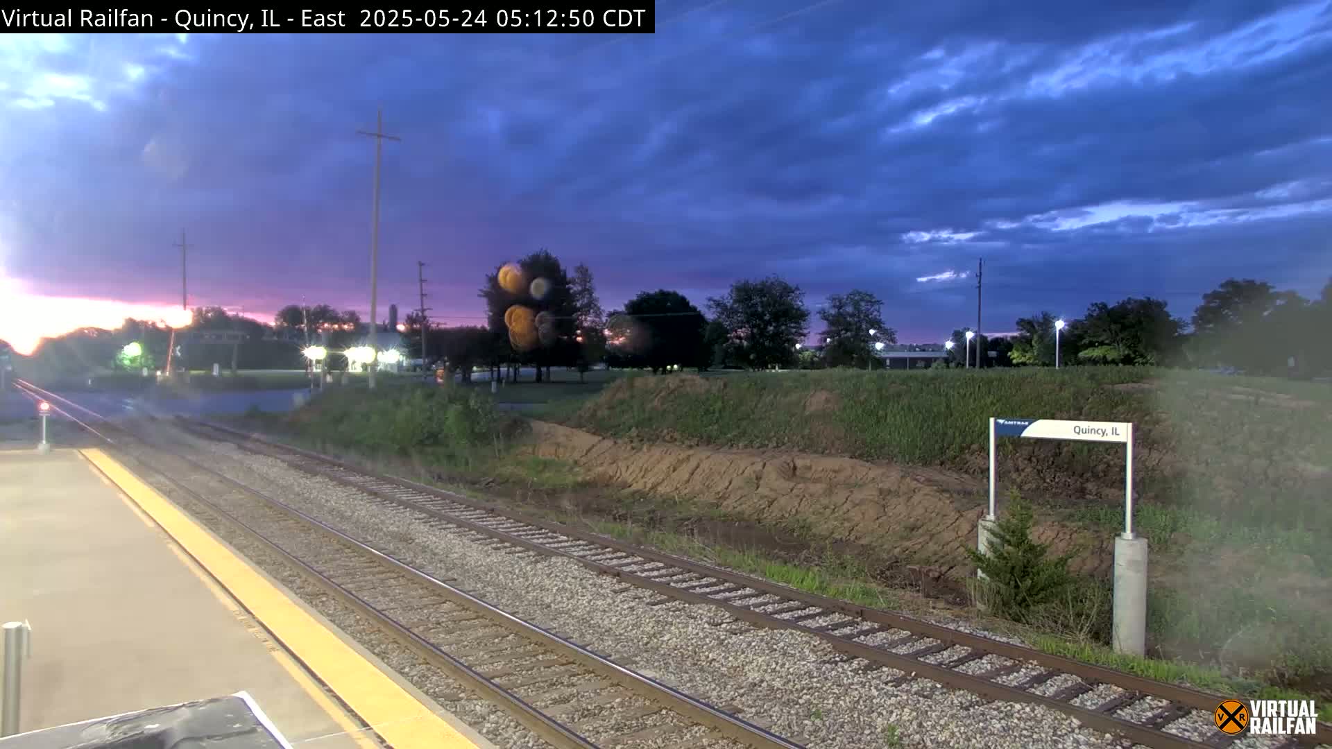 A train station platform at twilight under a mostly cloudy sky, with train tracks extending into the distance.