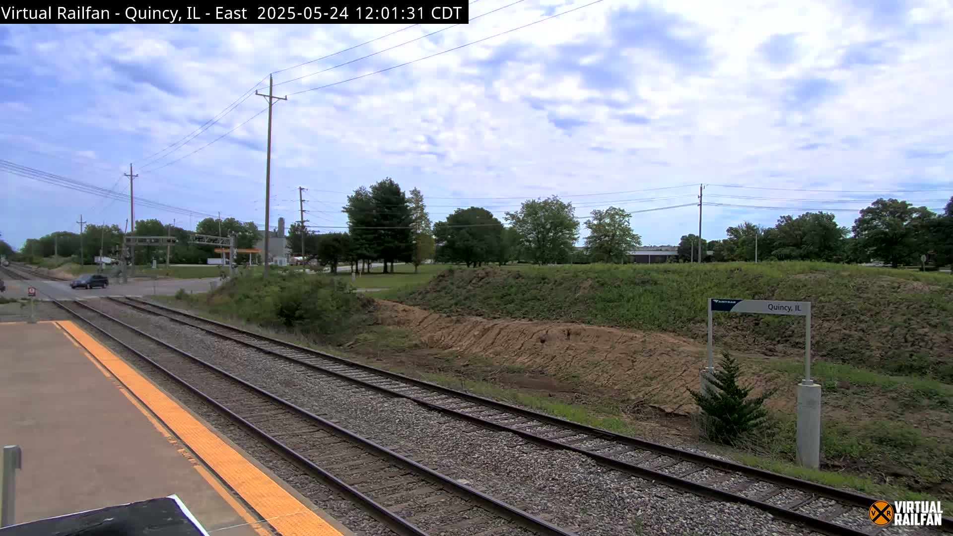 A view of train tracks at a station, with a road crossing in the background, under a partly cloudy sky.