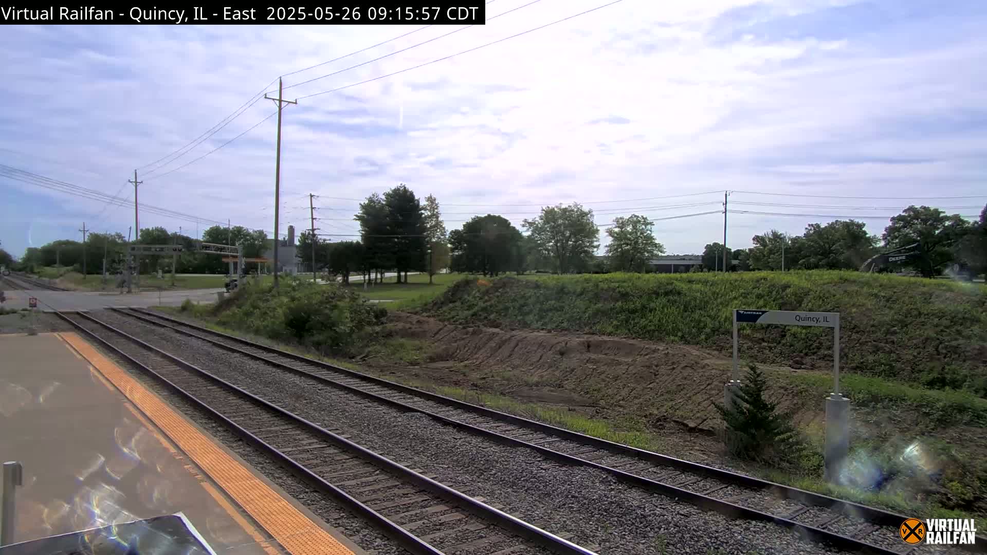 A view from a train platform shows several train tracks extending into the distance under a partly cloudy sky, with some greenery and earthworks visible beside the tracks.