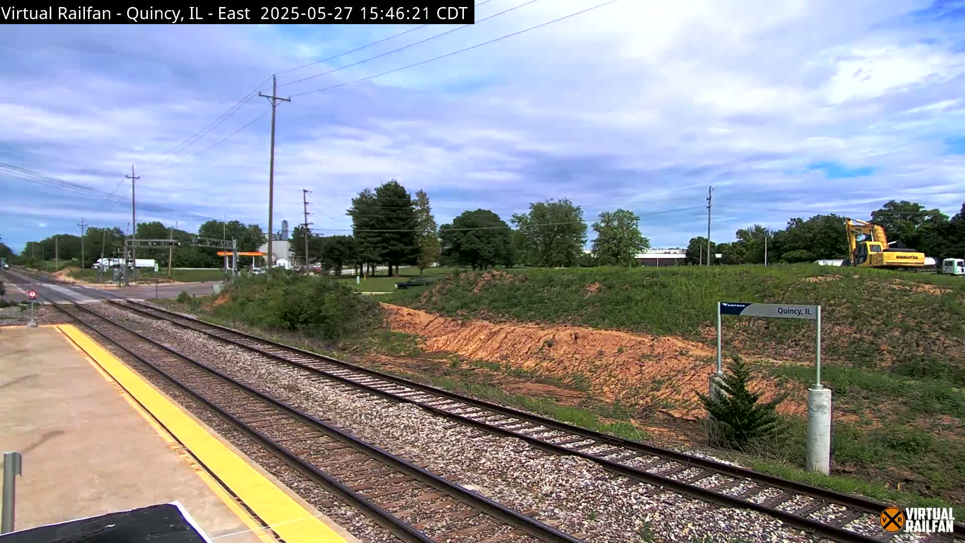 A partly cloudy day reveals a train platform next to railroad tracks, with a road crossing in the distance and some earthmoving equipment visible beyond a grassy hill.