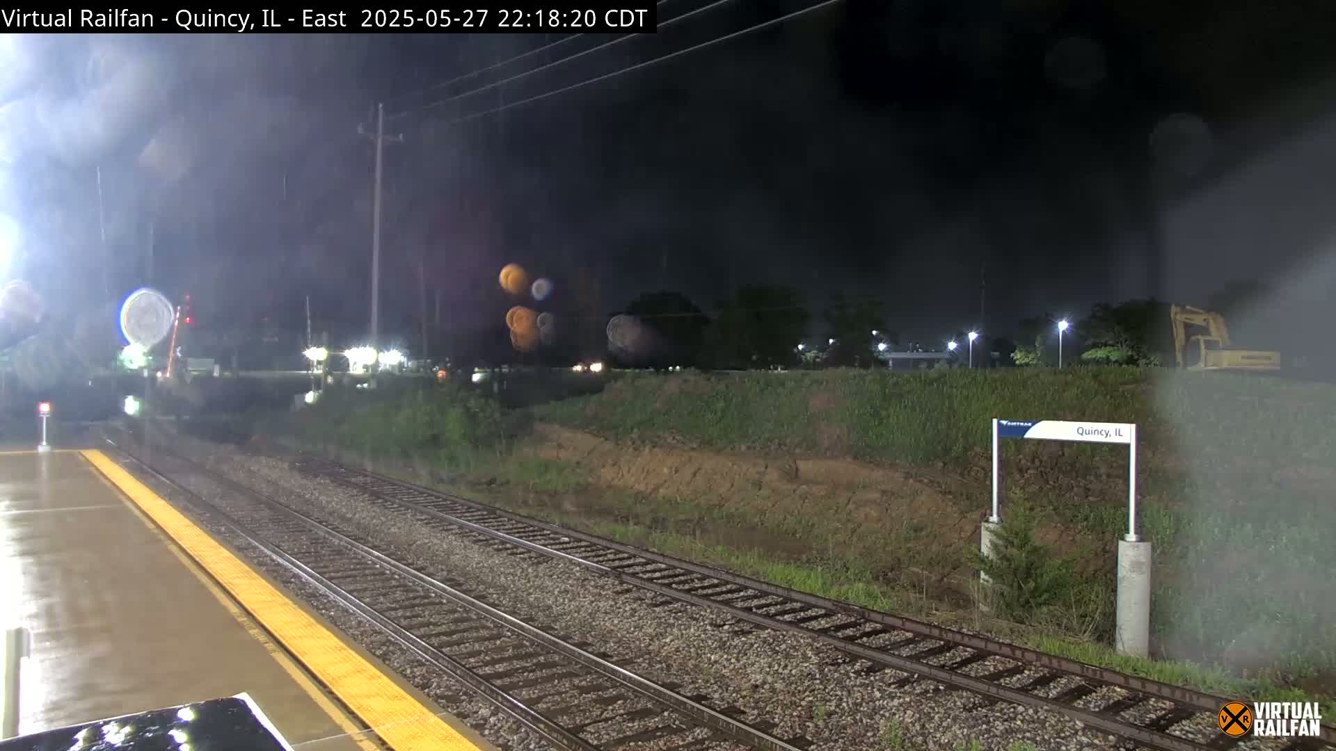 A nighttime view of a rain-slicked train platform and tracks, with a partially visible excavator in the background.