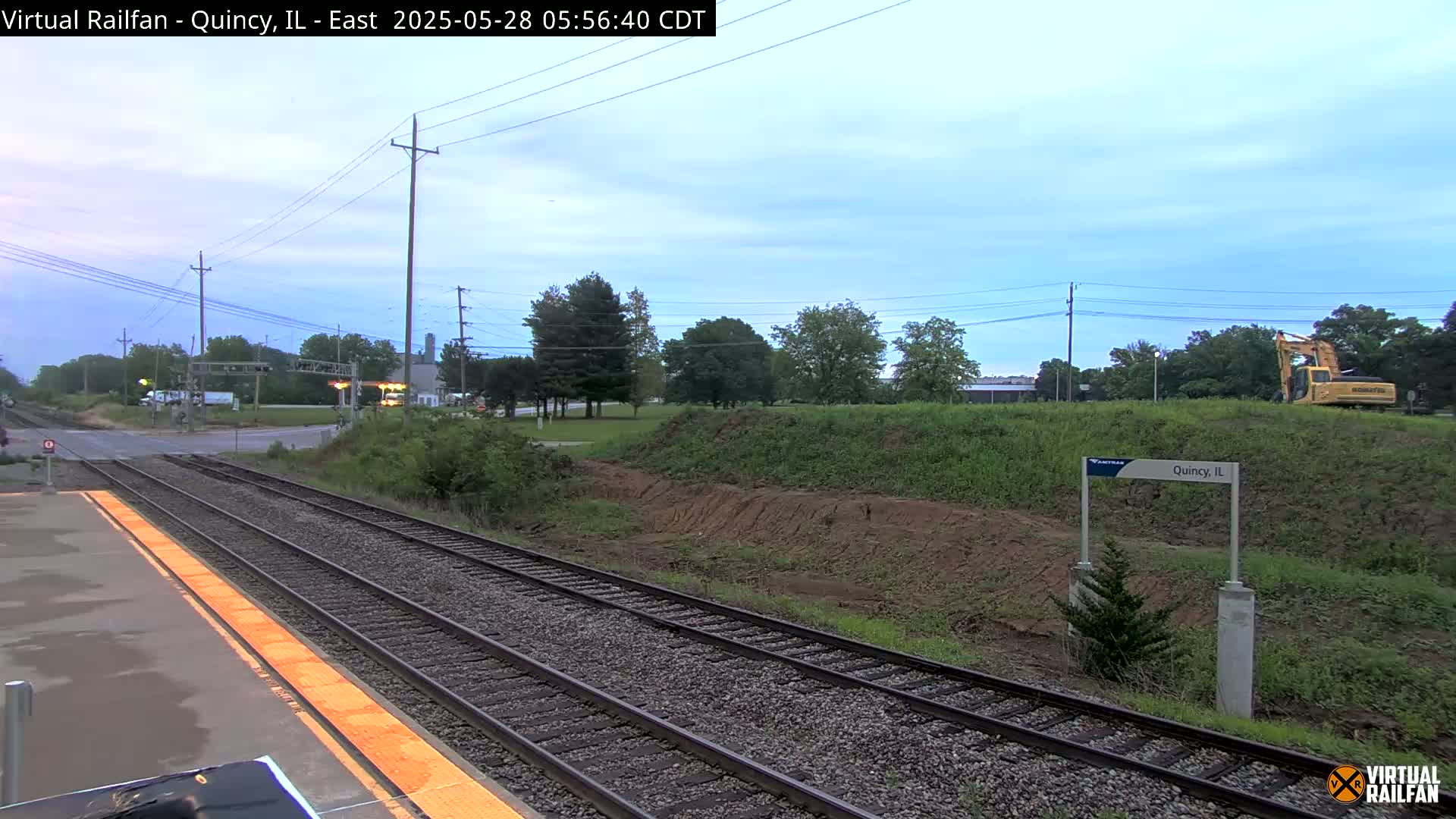 A train platform with multiple tracks under an overcast sky, showing construction activity on a nearby embankment with a yellow excavator visible.