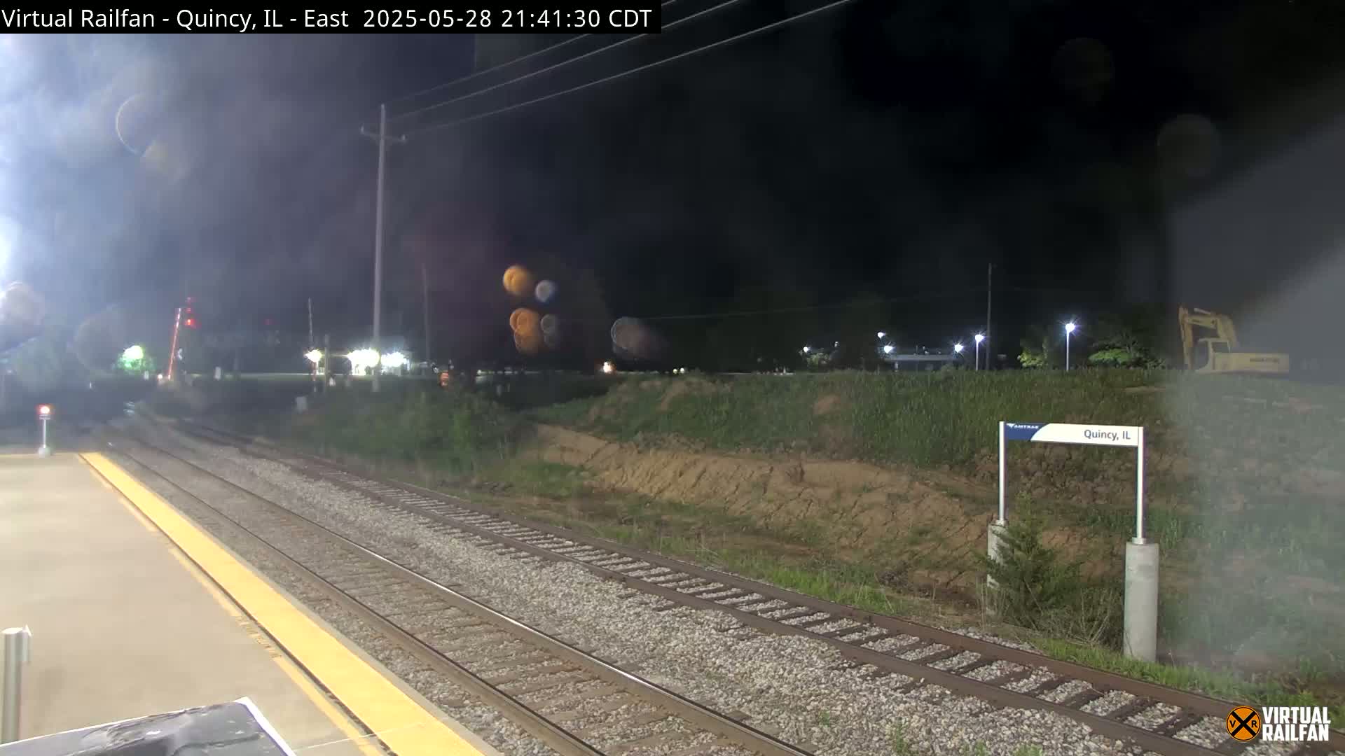 A nighttime view of train tracks at a station, with hazy conditions and some construction equipment visible in the distance.