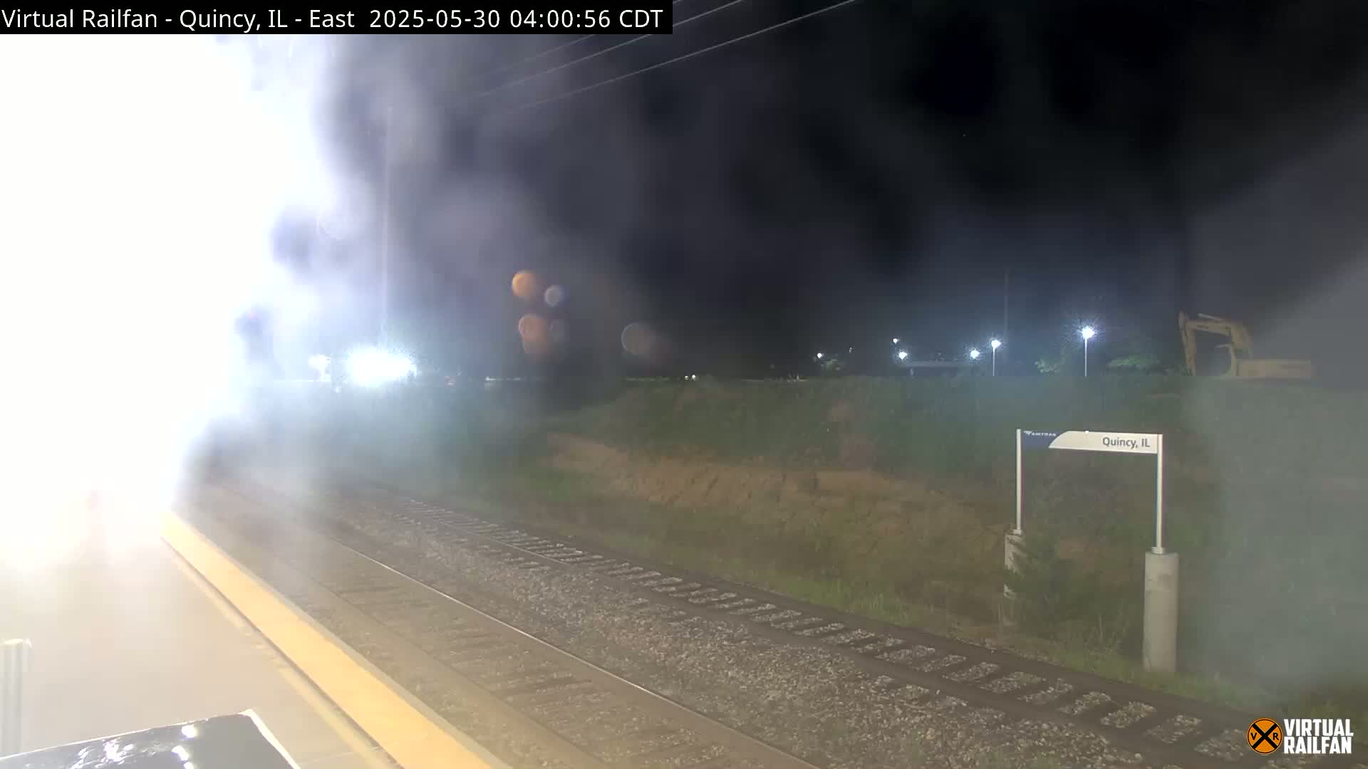 A nighttime view of a train platform with a train emitting steam, under hazy conditions.