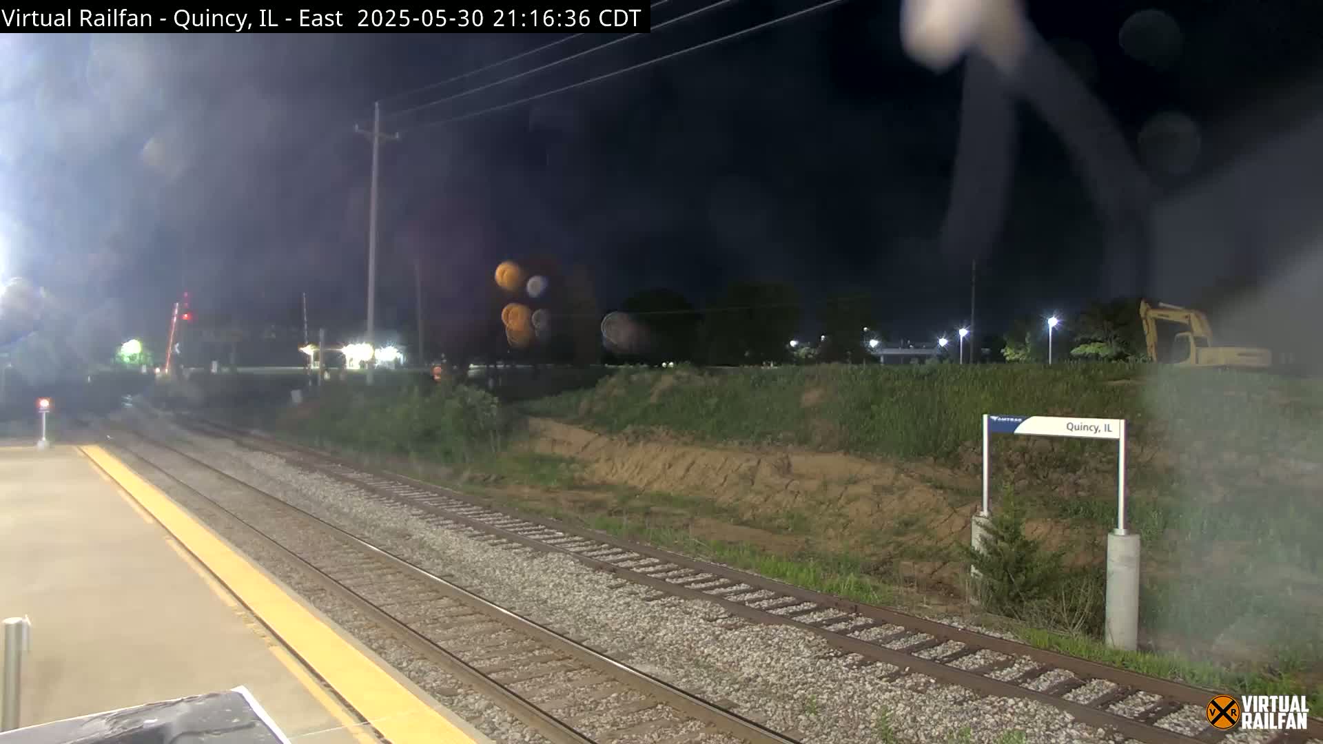 A nighttime view of a train platform with multiple train tracks, a small embankment, and a partially visible excavator in the background under hazy conditions.