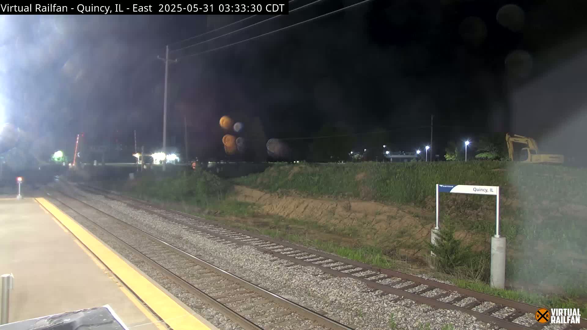 A nighttime view of train tracks at a station, with a small embankment and some construction equipment visible in the background under hazy conditions.
